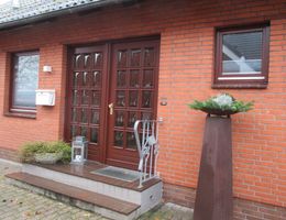 Außenansicht eines roten Backsteinhauses mit brauner Doppeltür, Fenstern, Briefkasten und dekorativen Pflanzgefäßen.
