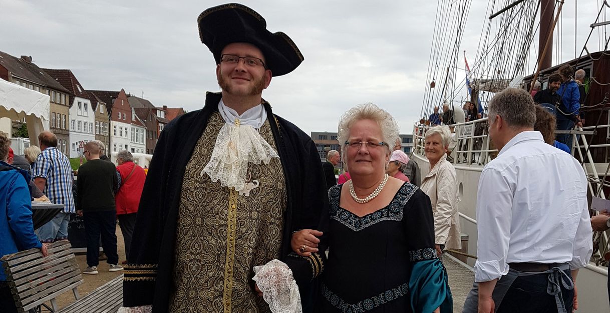 Ein Mann und eine Frau in historischen Kostümen stehen auf einem Hafenfest vor einem Segelschiff.