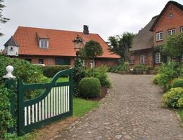 Ein gepflasterter Hof führt zu einem roten Backsteinhaus mit Ziegeldach; links ein grünes Tor, umgeben von üppigem Garten und Sträuchern.