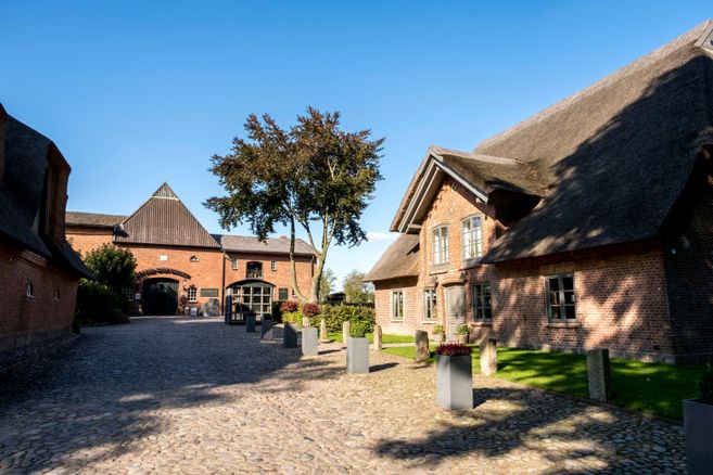 Ein gepflasterter Hof mit Backsteingebäuden und Reetdächern unter einem klaren blauen Himmel.