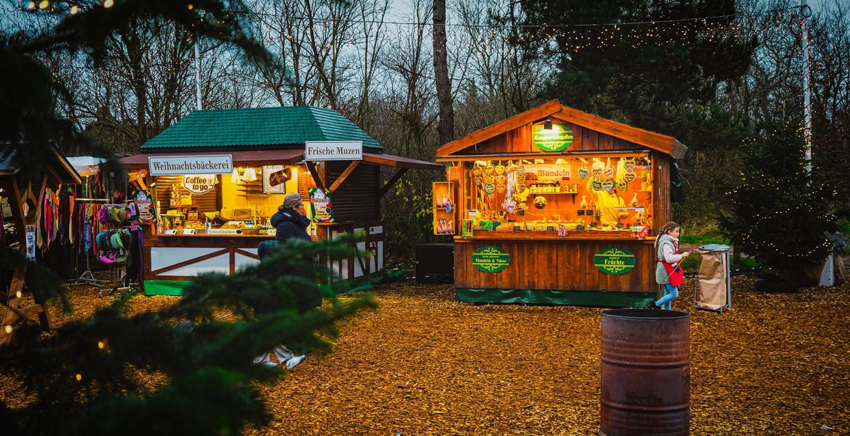 A Christmas market with wooden stalls outdoors. The booths offer various goods and foods, surrounded by trees and festive lighting.