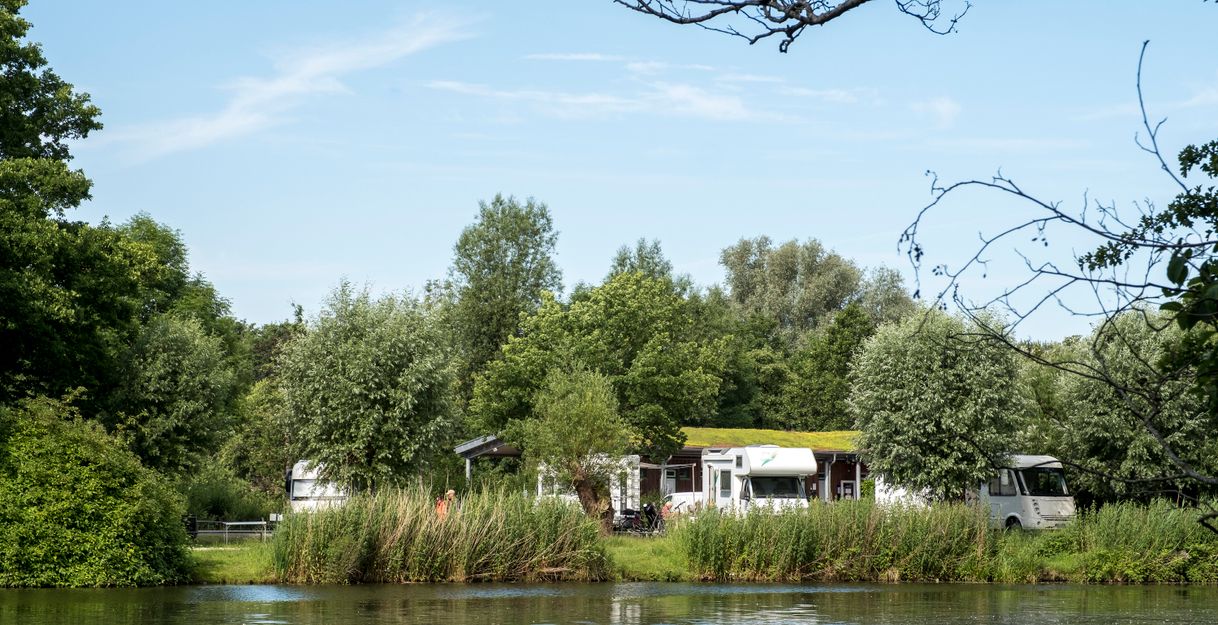 Wohnmobilstellplatz am Flussufer mit Bäumen und Wohnmobilen im Hintergrund.