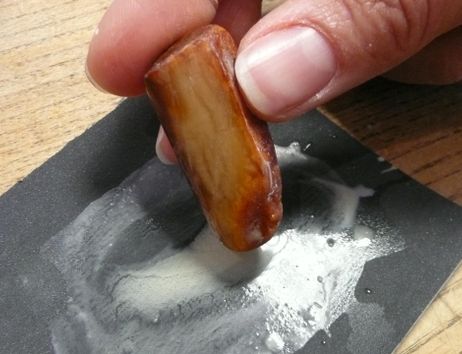 Ein Hand hält ein Stück Holz, das auf einem schwarzen Schleifpapier reibt. Es sind Spuren von Schleifstaub sichtbar.