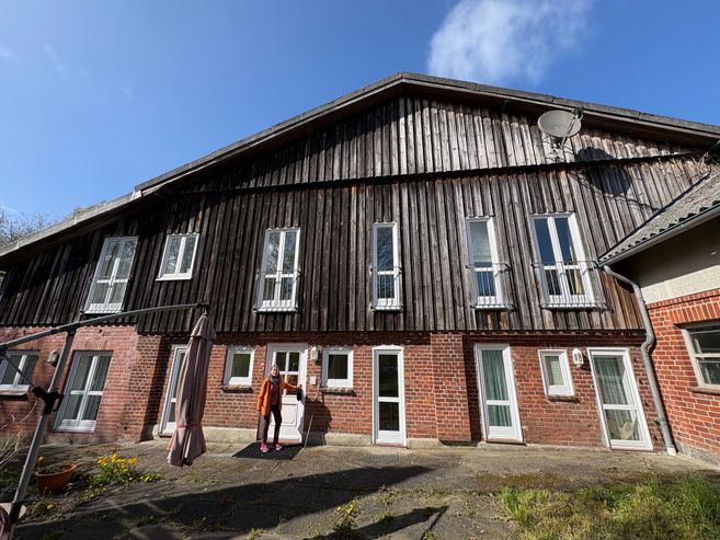 Ein zweistöckiges Gebäude mit einer Fassade aus Holz und Backstein. Eine Person steht vor der Eingangstür. Der Himmel ist blau und sonnig.