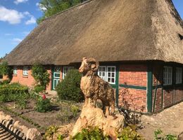 Eine Holzskulptur eines Schafbocks vor einem traditionellen Backsteinhaus mit Reetdach.