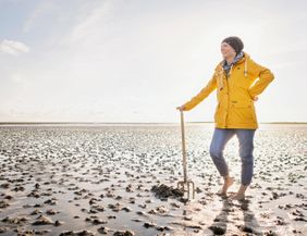 Eine Person in einem gelben Regenmantel steht barfuß am Watt. Im Hintergrund sind weite, schlickige Flächen und ein klarer Himmel zu sehen.