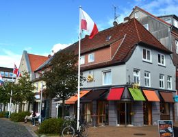 Außenansicht eines Hauses mit rotem Ziegeldach, bunten Markisen und mehreren Flaggen an einem Platz.